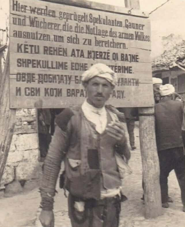 <br />
Në ballë të kësaj fotografie rri një shqiptar i moshës së mesme, i veshur me rroba të arrnuara me lecka e me tjerq galana, pantollona tradicional leshi për burra me ngjyrë të kafenjtë dhe shitira gajtani të zi, teksa me një cigare duhani në dorë, pozon në drejtim të objektivit fotografik. Më shumë sesa te vetë ai, vëmendja shkon mbrapa tij te një tabelë druri në sfond, në të cilën afërsisht i njëjti njoftim paralajmërues është shkruar në gjermanisht, shqip dhe serbisht. Teksti në gjermanisht ka këtë përmbajtje: “Këtu rrihen spekulantët dhe mashtruesit, si dhe fajdexhinjtë që shfrytëzojnë gjendjen e vështirë të njerëzve të varfër për t’u pasuruar”. Poshtë këtij teksti, në dy rreshta me shkronja të mëdha në shqip është shkruar: “Këtu rehën ata njerëz qi bajnë shpekullime dhe mashtërime”.